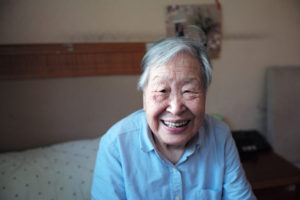 Elderly woman smiling broadly while sitting on bed in skilled nursing center.