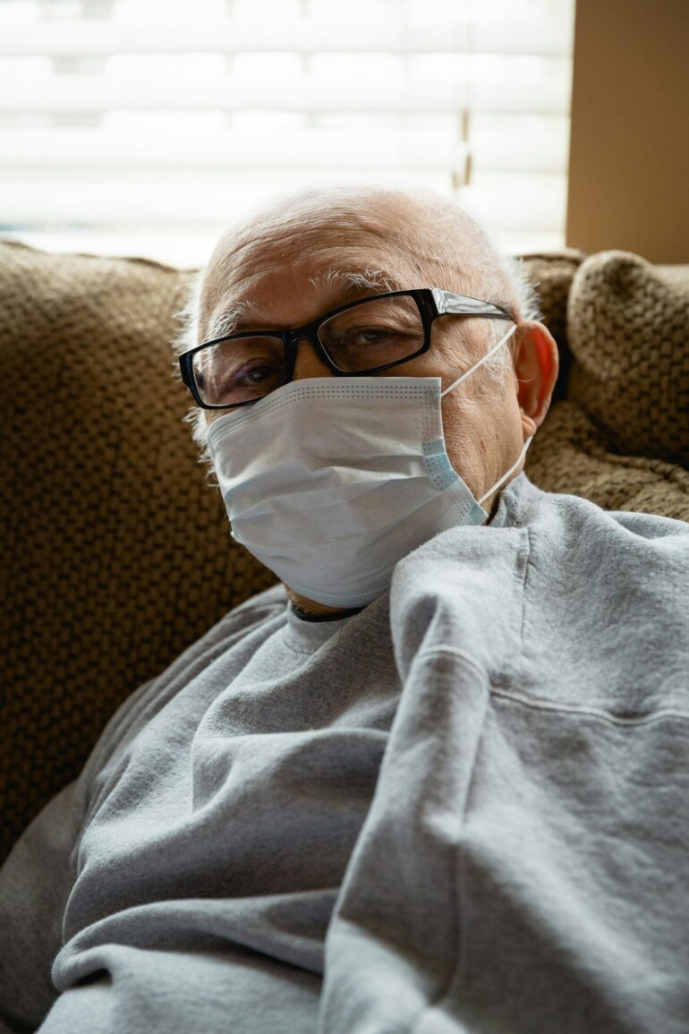 An elderly man wearing a mask to prevent infection, sitting on a couch.